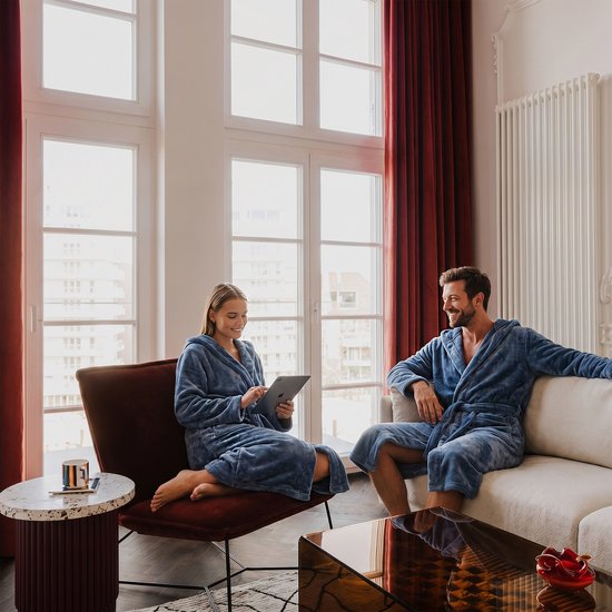 Homme et femme en peignoirs bleus se détendent dans un salon, regardant une tablette et l'un l'autre.
