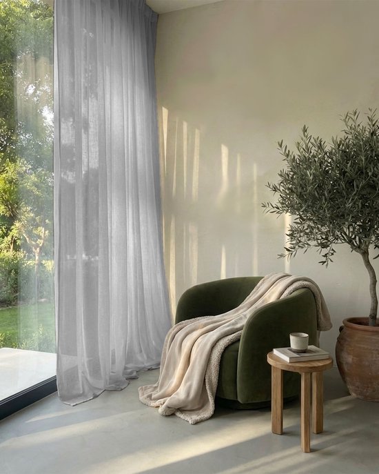 Een groene fauteuil met een beige plaid in een woonkamer met een transparant wit gordijn.