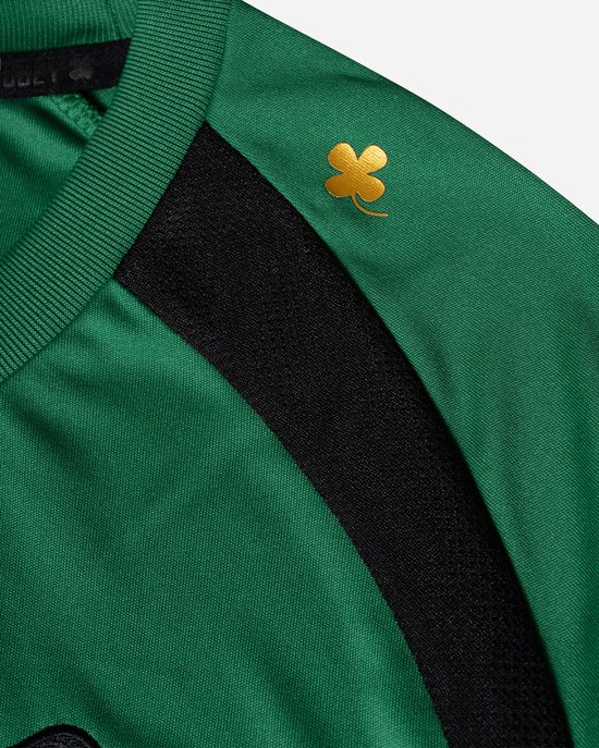 Detail van de rechterschouder van een groen sportshirt met zwarte accenten en een gouden klaver-applicatie.