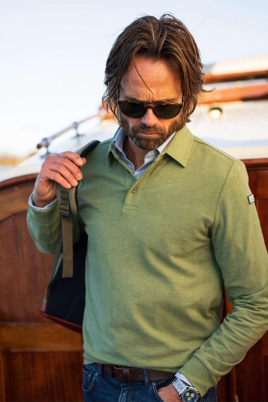Man met zonnebril die een lichtgroen poloshirt met lange mouwen draagt, deels over een schouder. Donkerblauwe jeans.