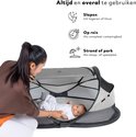 Moeder kijkt lachend naar baby in grijs-wit Deryan campingbedje op een kleed.