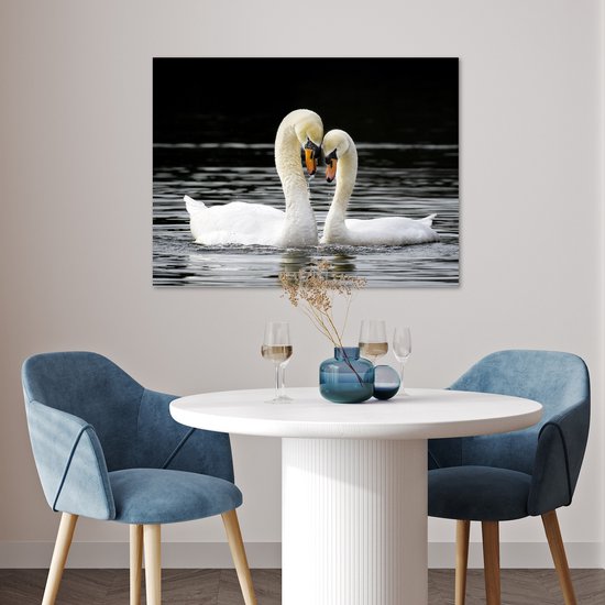 Deux cygnes blancs au bec orange nagent dans l'eau sombre, leurs cous forment un cœur, accrochés au mur au-dessus d'une table à manger.