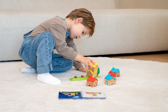 Een jongetje speelt met houten huisjes en blokken op een wit tapijt. Hij bouwt een toren met de blokken en plaatst huisjes naast de groene staaf.