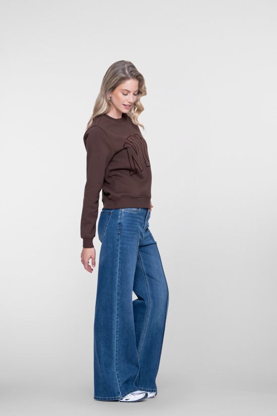 Vrouw draagt een donkerbruine sweater met 'NYC' opdruk en een wijde, stonewashed blauwe jeans. Ze staat opzij.