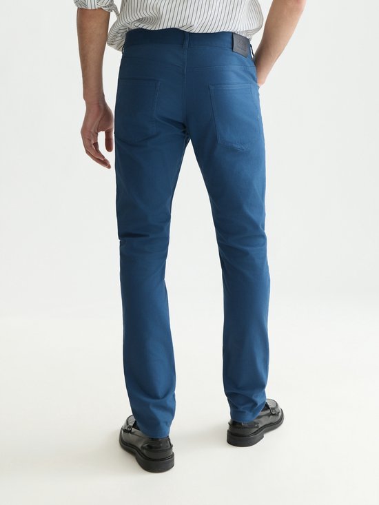 Model draagt een blauwe chino en gestreept overhemd, rugaanzicht met Scotch & Soda Amsterdam label.