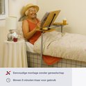 Lachende oudere vrouw in bed met strooien hoed, die leest op een verstelbare bedtafel met dienblad.