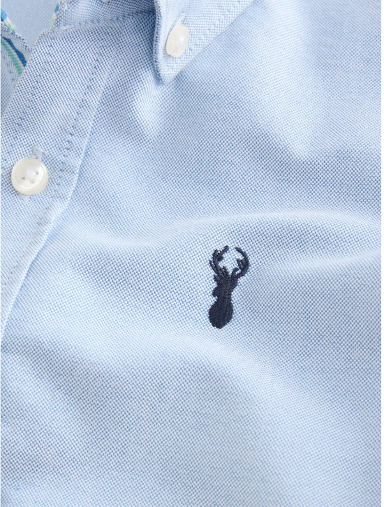 Close-up van het geborduurde donkerblauwe hert-logo op een lichtblauwe katoenen Oxford blouse.