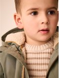 Portret van een jongetje in een groene parka over een gestreepte gebreide trui.