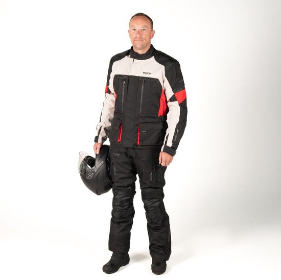 Man draagt een zwarte motorjas met lichtgrijze en rode accenten, en zwarte motorbroek. Houdt een helm vast.