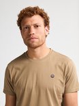 Close-up van een man met krullend rood haar, draagt lichtbruin t-shirt met borstlogo.