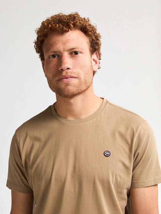 Close-up van een man met krullend rood haar, draagt lichtbruin t-shirt met borstlogo.