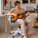 Een man speelt gitaar op een grijze bureaustoel met witte armleuningen en een wit onderstel, in een kamer met platenhoezen aan de muur.