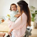 Lachende baby met lachrimpels wordt vastgehouden door moeder in roze geblokt shirt.