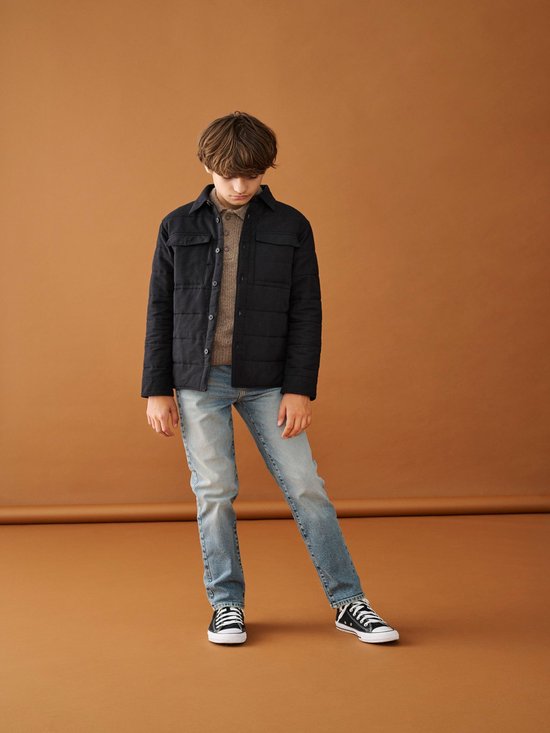 Jongens in een lichtblauwe jeans, bruin gebreid vest en een zwarte jas.