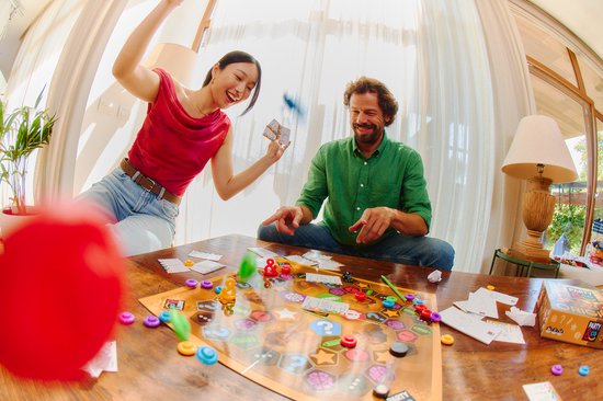 Een koppel, een vrouw met rode top en een man met groene blouse, spelen het spel Party & Co. De vrouw gooit kaarten omhoog, de man kijkt lachend toe.