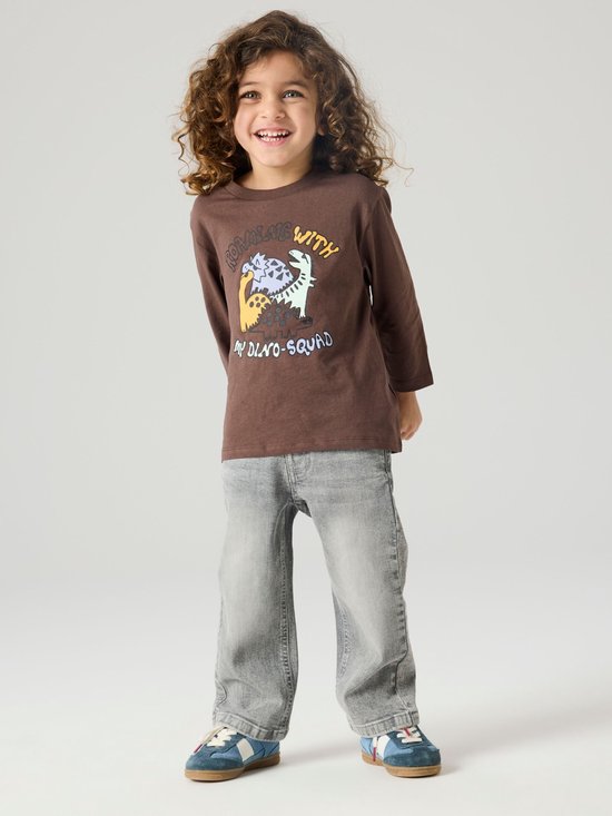 Jongen met krullend haar draagt een bruin T-shirt met dinosaurusprint en een grijze spijkerbroek.