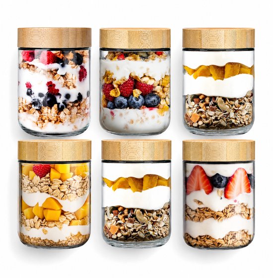 Zes glazen potten met bamboe deksel, gevuld met laagjes yoghurt, fruit en granola.
