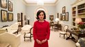 Natalie Portman in rode mantelpakje met parelketting, staat in salon.