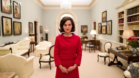 Natalie Portman in rode mantelpakje met parelketting, staat in salon.