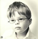 Een jongen met kort bruin haar en een bril, draagt een wit shirt. Zwart-wit foto.