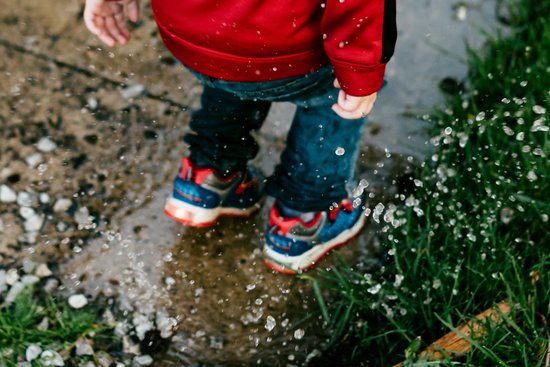Kind draagt blauwe sneakers met rode accenten en oranje zolen, spattend in plassen.