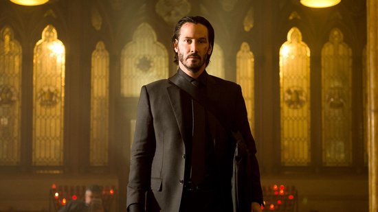 Keanu Reeves als John Wick, staand in een pak met een tas over zijn schouder.