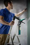 Man op een ladder gebruikt een wolfcraft kitpistool met groene en zwarte accenten om een muur af te werken.