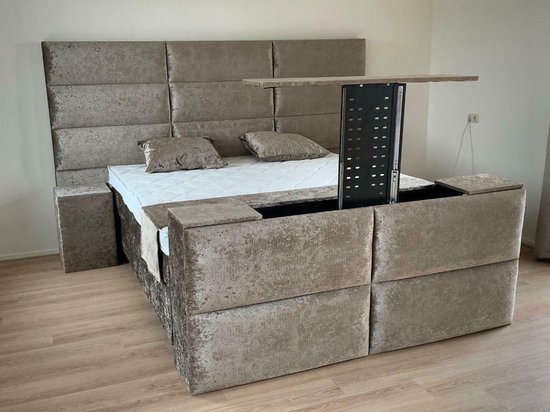 Grijze boxspring met TV-lift, met een gestoffeerd hoofdbord en kussens.