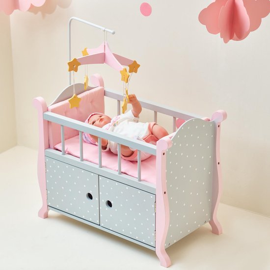 Roze poppenbedje met grijze zijkanten met witte stippen en twee opberglades aan de voorkant. Een baby in een wit pakje ligt erin. Met sterrenmobiel.