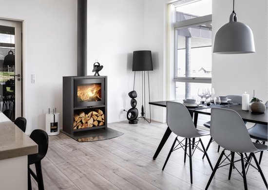 Moderne woonkamer met een brandende houtkachel, naast een eettafel met stoelen. Een zwarte kachelventilator staat op de kachel.