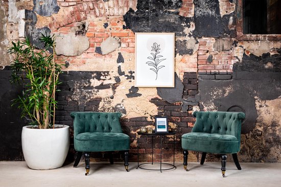 Deux fauteuils en velours vert émeraude capitonné, devant un mur de briques noires, avec une plante et une œuvre d'art murale.