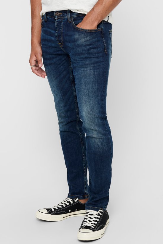 Man in donkerblauwe skinny jeans met lichte slijtage en zwarte sneakers met witte veters.