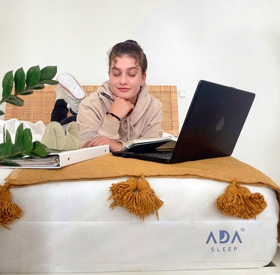 Jonge vrouw ligt op haar buik op een matras en werkt op een laptop. Een deken met franjes ligt over de matras.