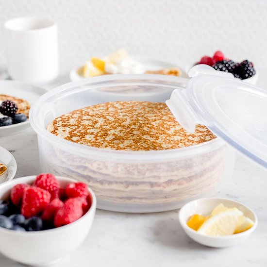 Pile de crêpes dans une boîte de conservation ronde et transparente. À côté de la boîte se trouvent des bols de baies et de citron.