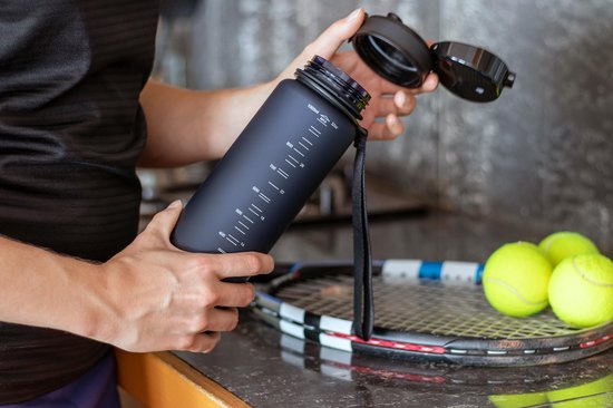 Persoon houdt matzwarte King Mungo drinkfles met schaalverdeling vast, bij tennisrackets en ballen.