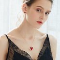 Een model draagt een gouden ketting met een rode hartvormige hanger met vleugels en een bijpassende oorbel.