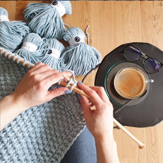 Blauwe Bobbiny macramé garenbundels en handen die breien met houten naalden.