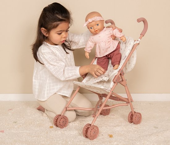 Een kind in witte blouse kleedt een pop aan met roze kleding in een roze poppenbuggy met bloemenpatroon.