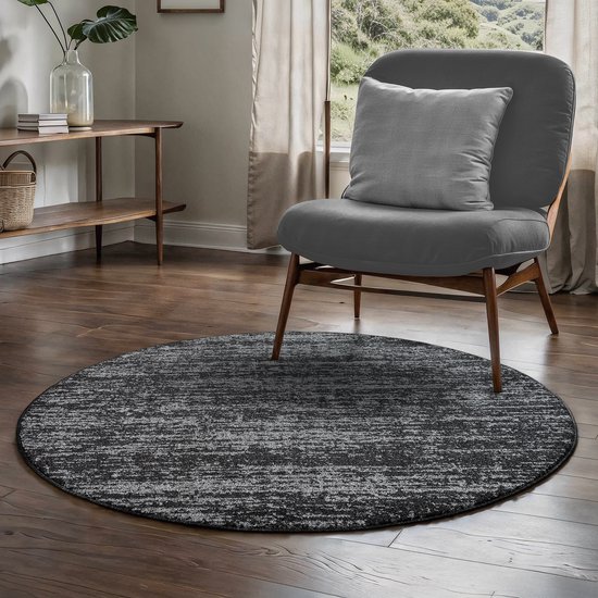 Rond zwartgrijs vloerkleed met een modern, gemêleerd patroon, geplaatst voor een grijze stoel in een woonkamer.