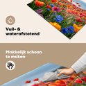 Een close-up van een kookplaatafdekking met rode, oranje, roze en blauwe klaprozen. Een hand veegt het oppervlak schoon.