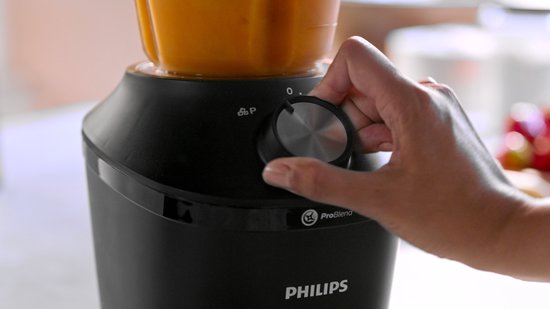 Hand draait aan de knop van een zwarte Philips blender met oranje vloeistof.