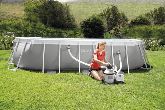 Vrouw onderhoudt een Intex zwembadfilterpomp naast een groot Intex zwembad in een tuin.