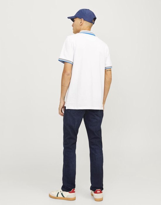 Man draagt een donkerblauwe jeans en een witte polo met blauwe kraag en mouwranden, met blauwe pet.