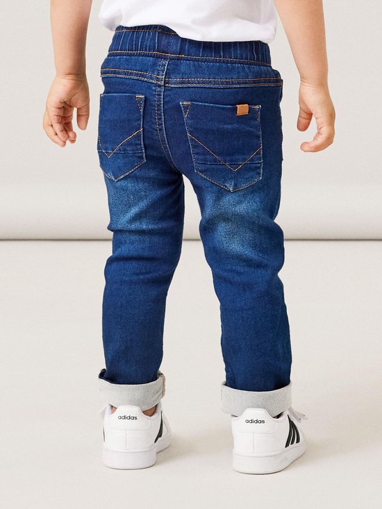 Achterkant van een jongetje in een donkerblauwe spijkerbroek met stonewashed effect, trekkoord en oranje stiksels. Omgeslagen broekspijpen.
