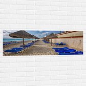 WallClassics - Muursticker - Blauwe Ligbedden op Strand met Rieten Parasols - 120x40 cm Foto op Muursticker