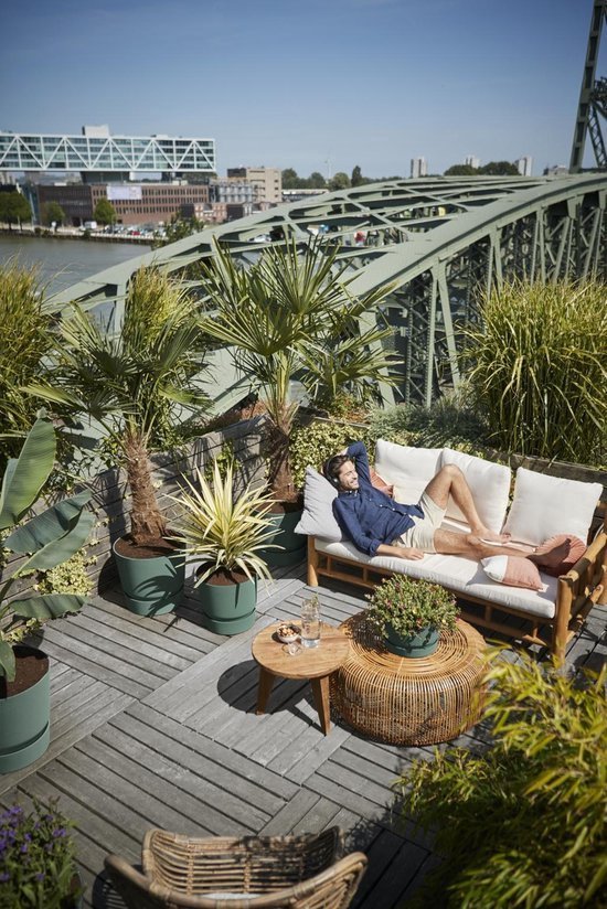 Man ontspant op een loungebank op een houten terras, omringd door palmbomen en planten in olijfgroene Elho potten.