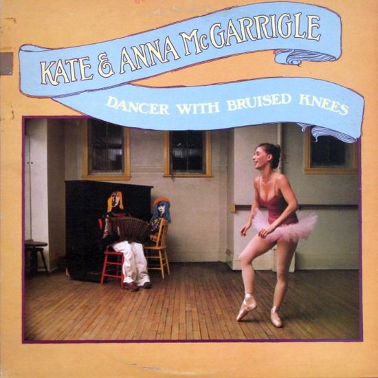 Een ballerina in een roze tutu en rode top danst op spitzen. Twee poppen zitten op stoelen op de achtergrond.