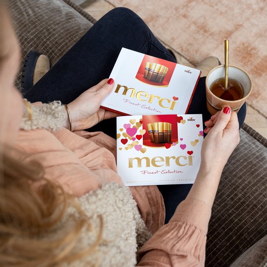 Een persoon houdt twee Merci dozen vast. De dozen hebben een roze en rode hartjesprint en 'Merci Finest Selection' in gouden letters.