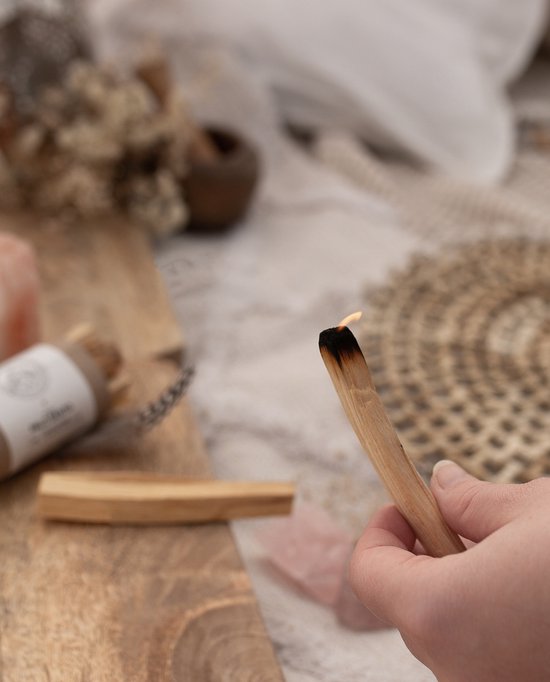 Een hand houdt een brandende Palo Santo stok vast, met onverbrande stokjes en roze kristallen op de achtergrond.