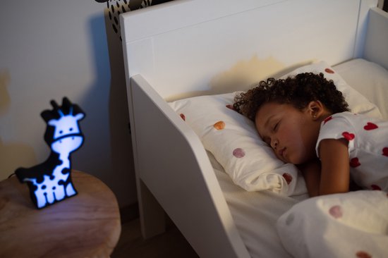 Wit oplichtend giraf-nachtlampje naast een slapend meisje in een bed.